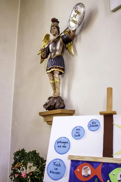 Geflügelte Michaelsfigur in Rüstung mit Schild, den Fuß auf eine dunkelhäutige Teufelsgestalt gesetzt, davor eine Kinder-Pinnwand