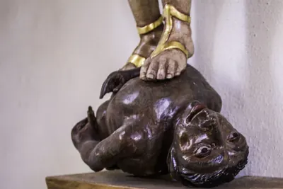Dunkelhäutige Teufelsgestalt mit Krallenhänden, die rücklings unter dem Fuß einer Heiligenfigur am Boden liegt