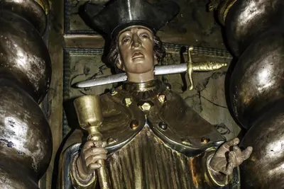 Skulptur eines Geistlichen mit Birett, dem ein Schwert quer durch den Hals getrieben ist, in der Hand ein Kelch