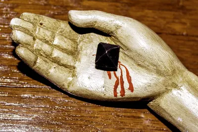 Hand einer Kruzifixfigur mit eingeschlagenem Nagel, aus der Wunde laufen rote Blutspuren