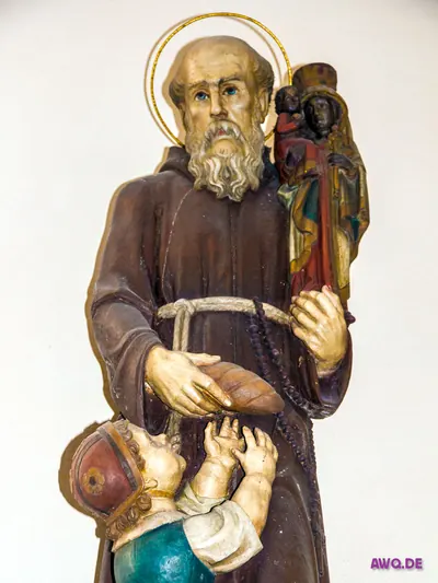 Bemalte Figur eines Ordensmannes mit Heiligenschein, Rosenkranz und schwarzer Madonna auf der Schulter, der einem Kind ein Brot reicht