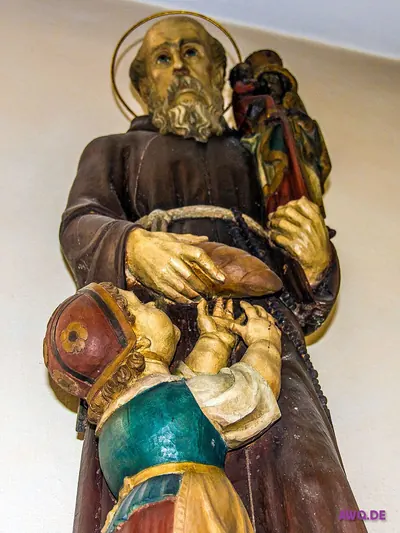 Untersicht auf eine Ordensmannfigur mit schwarzer Madonna auf der Schulter; ein Kind greift nach dem Brot in ihren Händen
