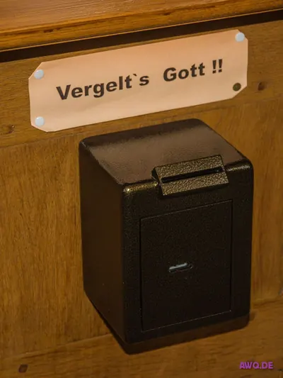 Schwarzer Opferstock an einer Holzbank, darüber ein Schild mit der Aufschrift „Vergelt’s Gott!!“