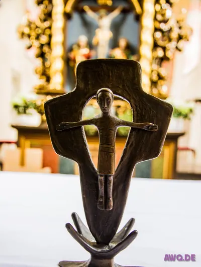 Modernes Bronzekreuz mit stilisierter Christusfigur auf dem Altartisch, dahinter ein barocker Hochaltar
