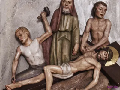 Kreuzwegrelief: Männer nageln den am Boden liegenden Jesus ans Kreuz, einer holt mit dem Hammer aus