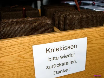 Kniekissen in einem Kirchenbankfach, darauf ein Schild „Kniekissen bitte wieder zurückstellen. Danke!“