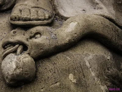 Verwittertes Steinrelief: ein Fuß tritt auf den Kopf eines gehörnten Drachen, der eine Kugel im Maul hält