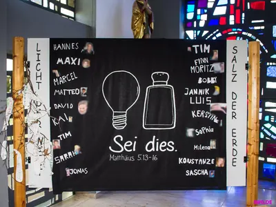 Schwarze Stellwand mit Glühbirne und Salzstreuer, Aufschrift „Sei dies.“ und vielen Vornamen