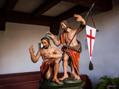 Figurengruppe der Taufe Jesu: Johannes gießt Wasser über den knienden Christus, daneben eine Kreuzfahne