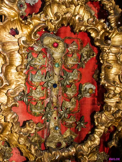 Reliquiar: ein mit Goldfäden und Schmucksteinen verzierter Knochen auf rotem Samt