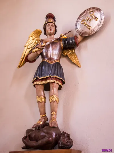 Michaelsfigur in Rüstung mit Schild „Quis ut Deus“, unter ihren Füßen eine dunkelhäutige Teufelsgestalt