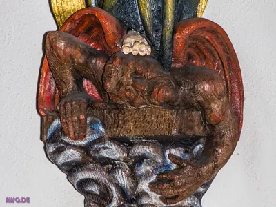 Bemaltes Holzrelief: eine dunkle, geflügelte Gestalt liegt über einem Balken, darunter weiß-blaue Wellen