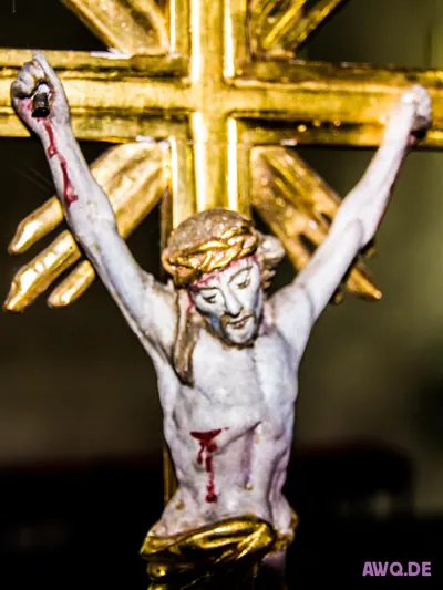 Kleine weiße Kruzifixfigur mit blutenden Wunden vor einem goldenen Strahlenkreuz