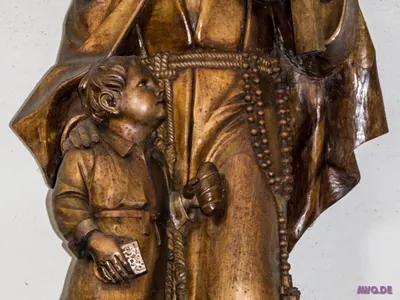 Detail einer Holzfigur: ein Kind mit einem Heftchen in der Hand blickt nach oben
