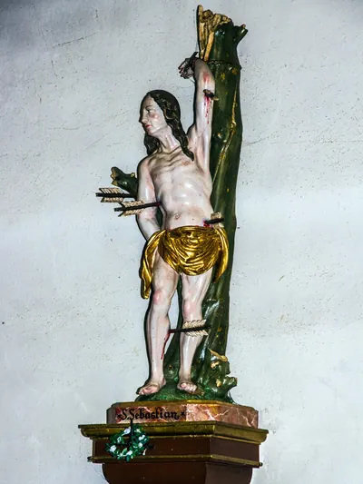 Sebastiansfigur am grünen Baumstamm, von Pfeilen durchbohrt, auf dem Sockel der Name S. Sebastian