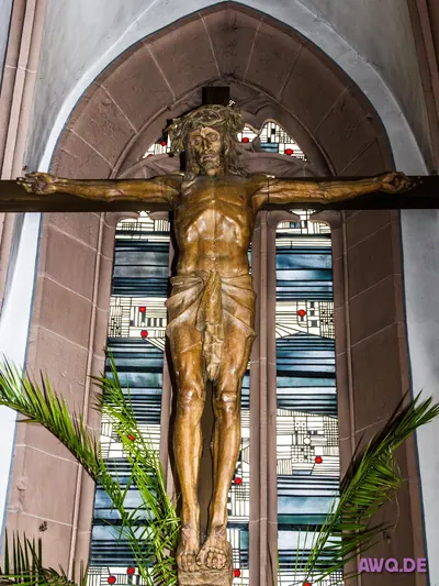 Großes Holzkruzifix vor bunten Kirchenfenstern, davor Palmwedel